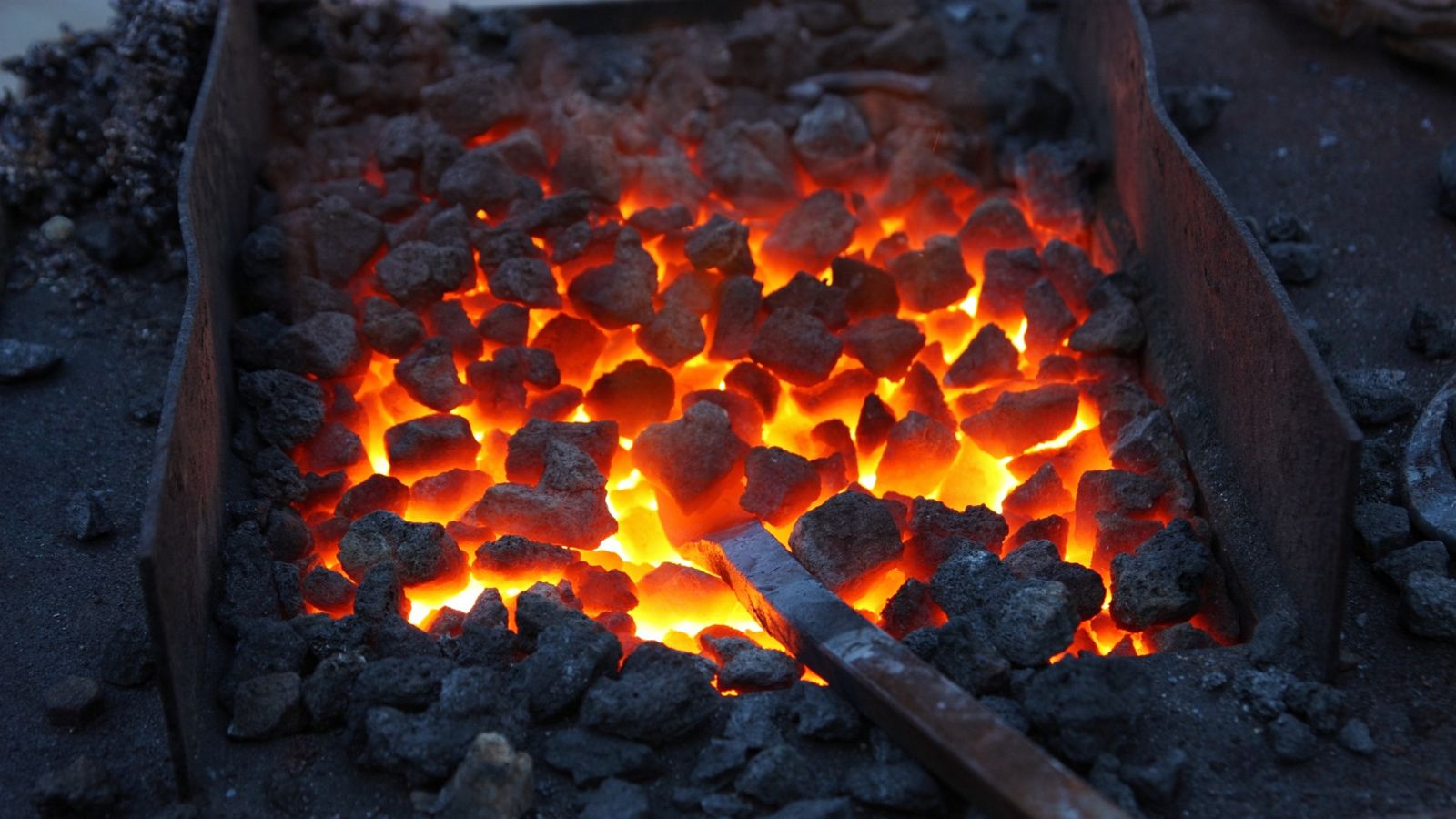 Close-up of glowing red-hot forge coals in an open hearth with a steel tool embedded — heat treatment as atom-time, the kind of physical process that cannot be sprinted.