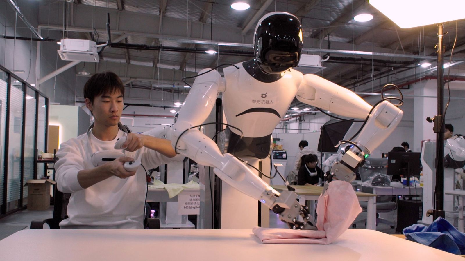 A humanoid robot folding cloth on a lab bench, teleoperated by an engineer holding motion controllers