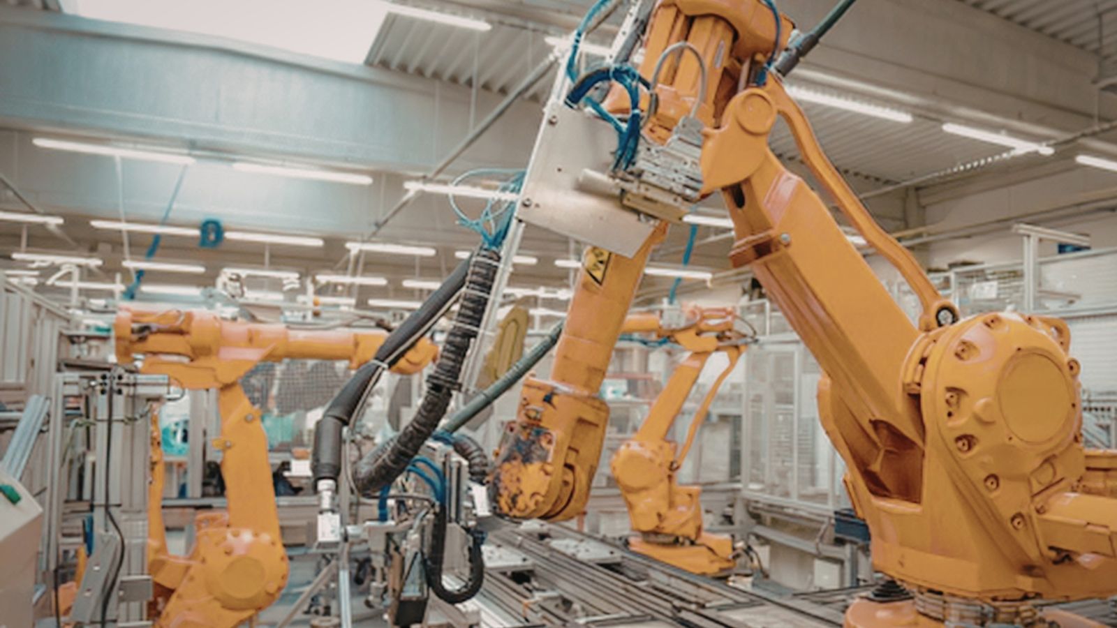 A cluster of orange industrial robot arms over a shop-floor cell with overhead lighting, depth into the production hall