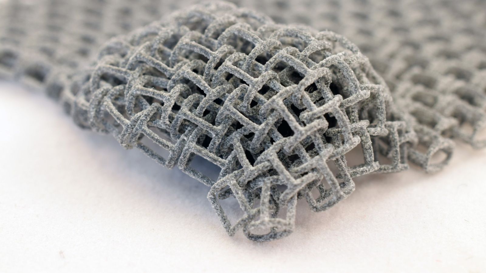 Macro of a metal additive-manufactured lattice — woven cube cells in sintered grey, the literal artefact of qualification at the geometry-and-powder level.