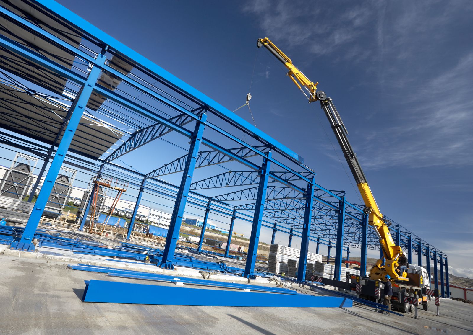 Blue steel frame of an industrial building mid-construction, a yellow mobile crane lifting a beam against a clear sky — proximity being built one lift at a time