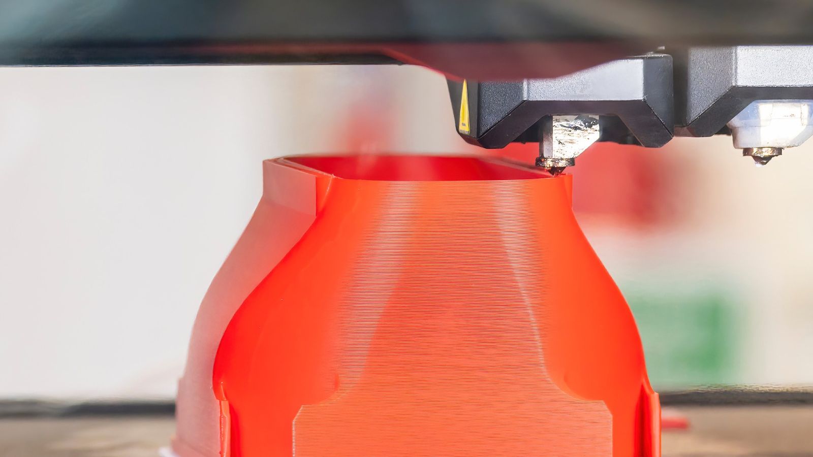 Close-up of an FDM printer extruder mid-build over a red plastic form on the build plate