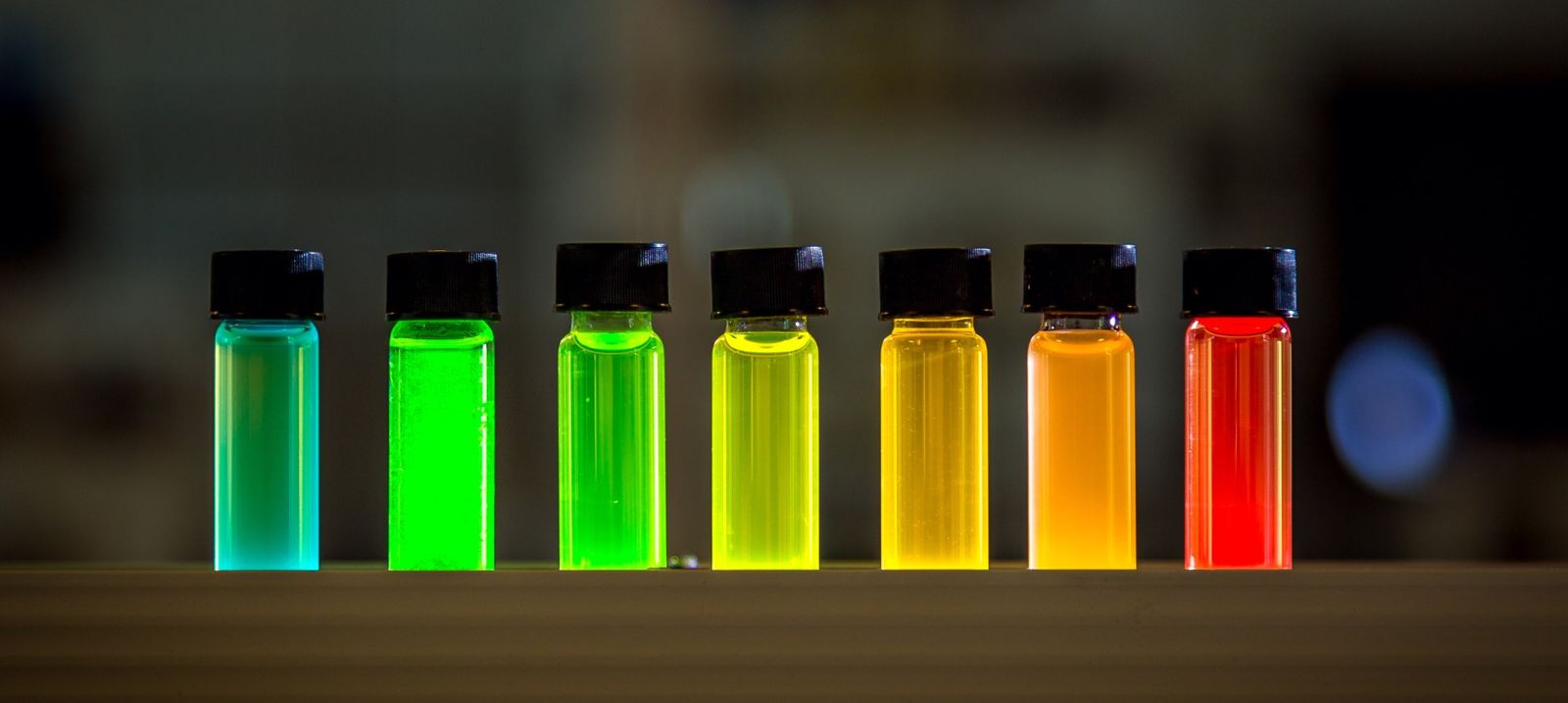 A row of small glass vials with brightly-coloured liquids in cyan, green, yellow, orange and red on a dark surface — quantum-dot synthesis, the kind of deep-tech IP that sits in the supply chain where value concentrates and sovereignty resides.