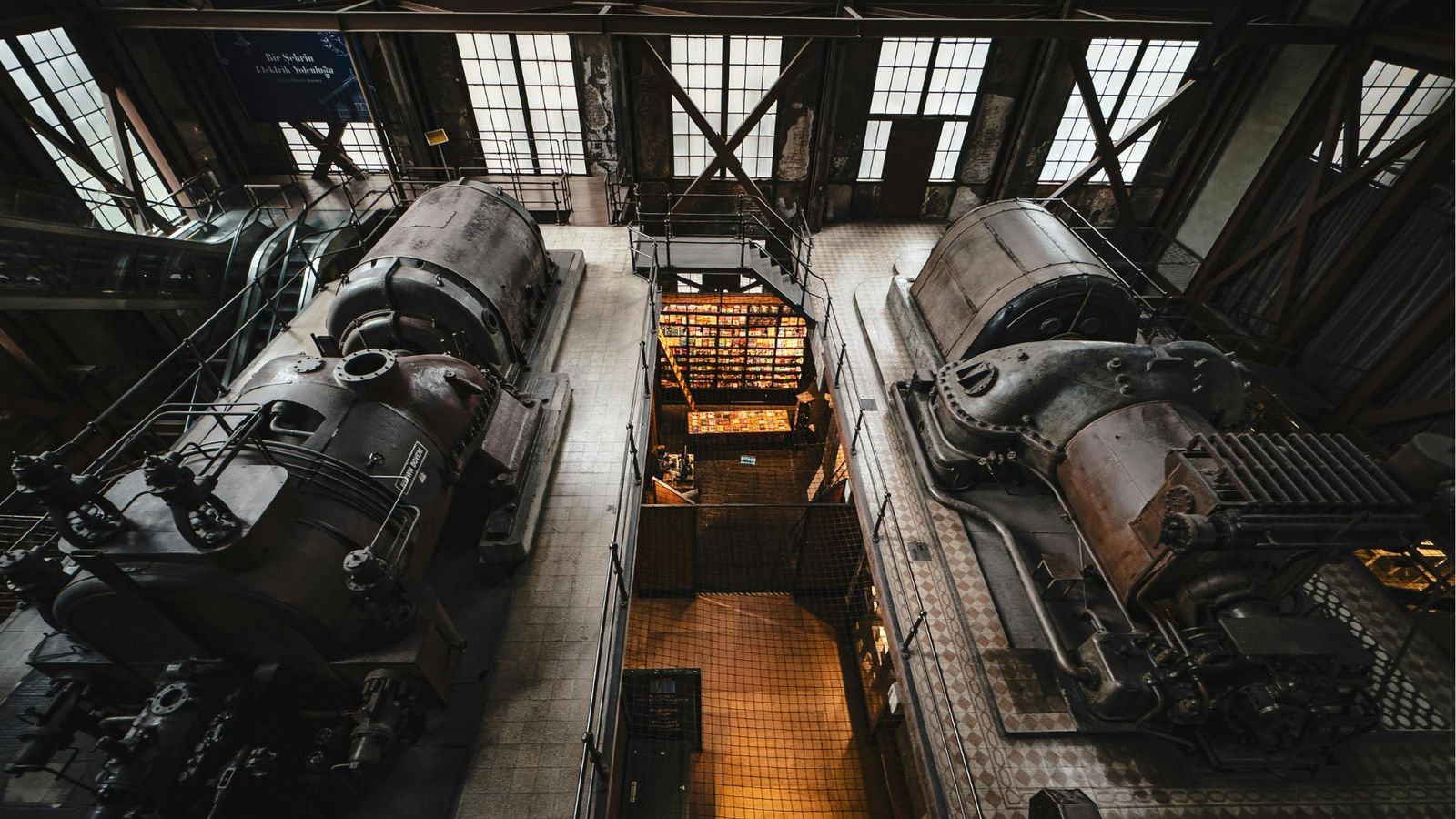 Two huge heritage industrial engines facing each other in a preserved engine hall, daylight pouring through arched factory windows
