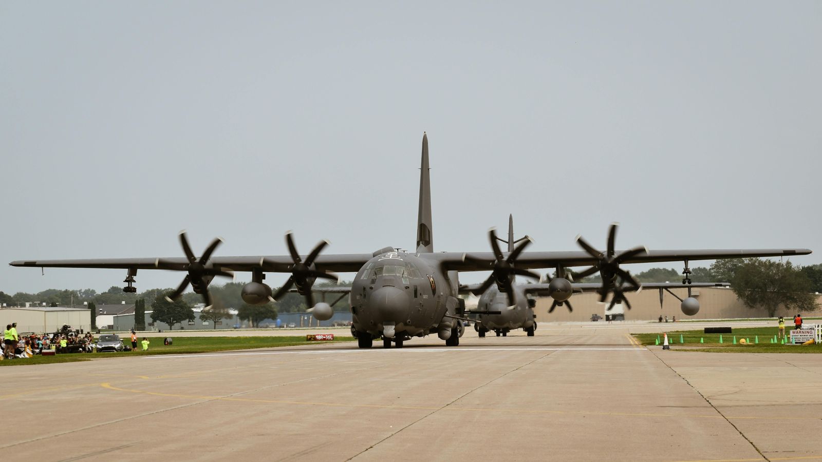 A four-engine military transport aircraft taxiing head-on down an apron, propellers turning, under flat grey daylight