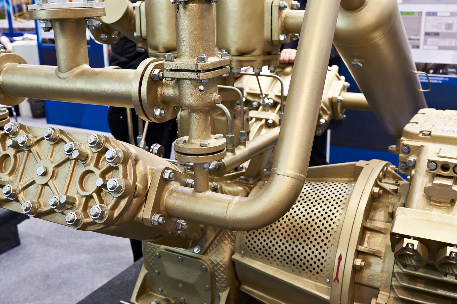 Large brass-finished pump and pipework assembly on a trade-show stand, flanges, bolts and a perforated guard in close detail under exhibition lighting