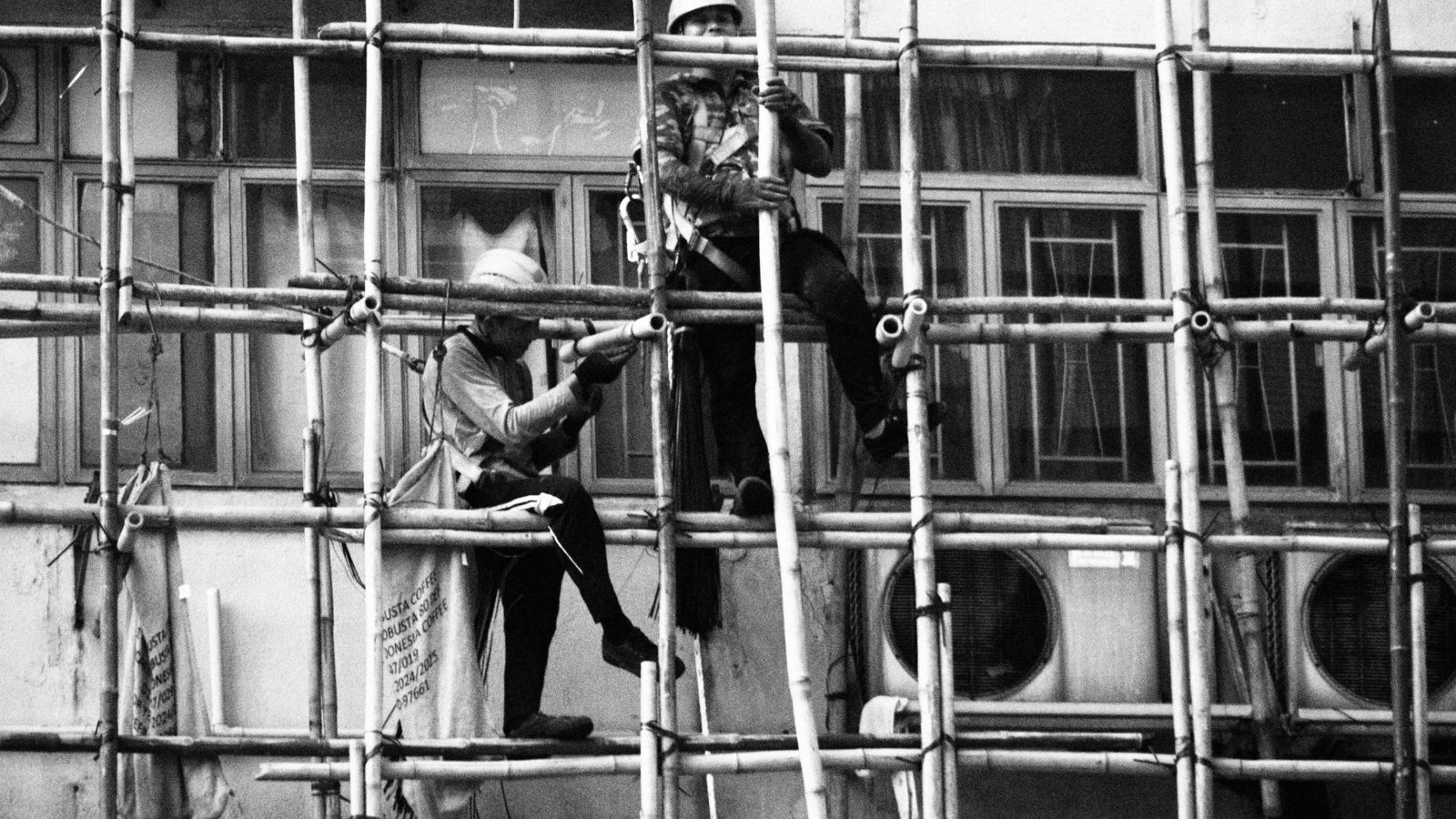 Two workers on bamboo scaffolding against a tiled facade, the poles and rungs at uneven heights, black and white