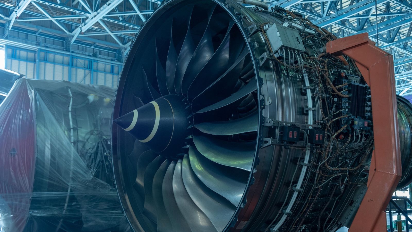 Large turbofan engine opened up in a maintenance hangar, fan blades in the foreground and harness looms behind