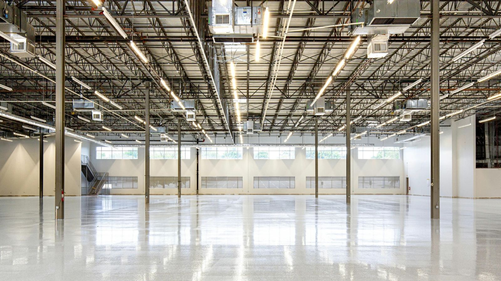 Pristine empty manufacturing facility interior with polished epoxy floor, exposed roof truss with linear lighting, and clean white-clad walls — a funded shell with nothing producing in it